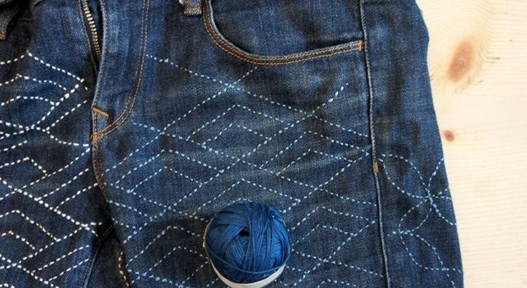 Närbild av jeans med synliga vita sashikostygn och blå trådrulle